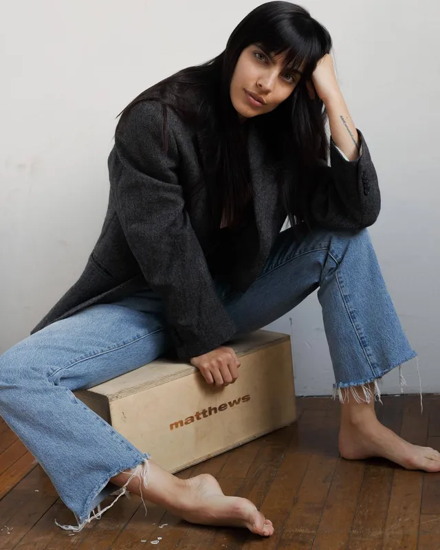 María Gabriela de Faría sitting barefoot in jeans beside a wooden box.