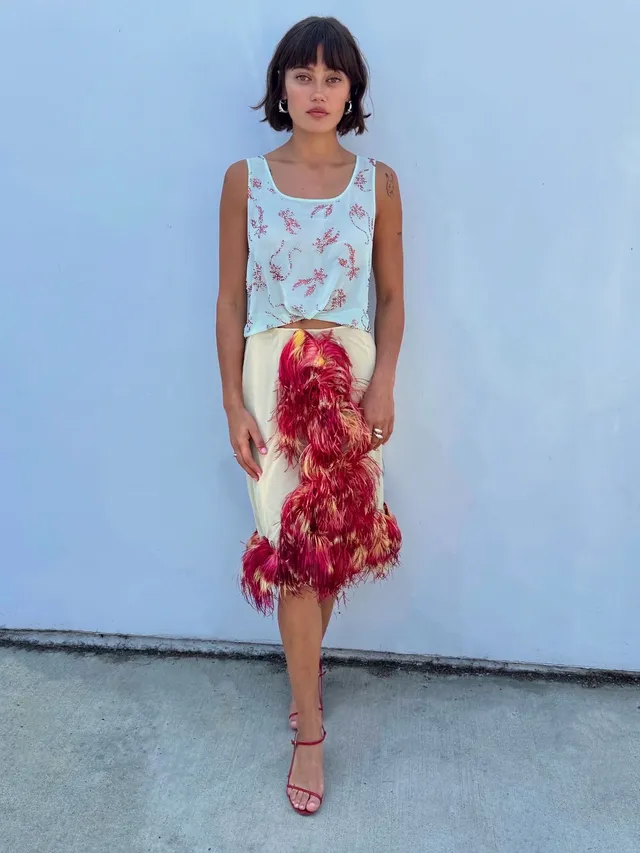Ella Purnell standing against a pale wall in a feather trimmed skirt.
