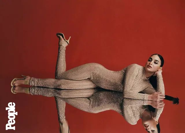 Melissa Barrera reclining in a gold outfit and heels on a reflective floor.