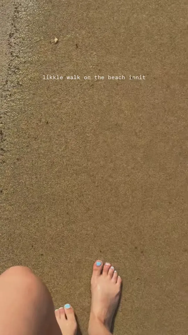 Rachel Zegler standing barefoot on wet sand beside the sea.