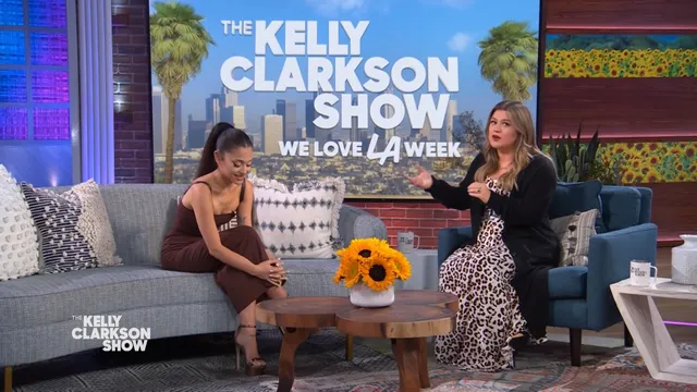 Ariana Grande seated on a television set in a dark dress and heels.