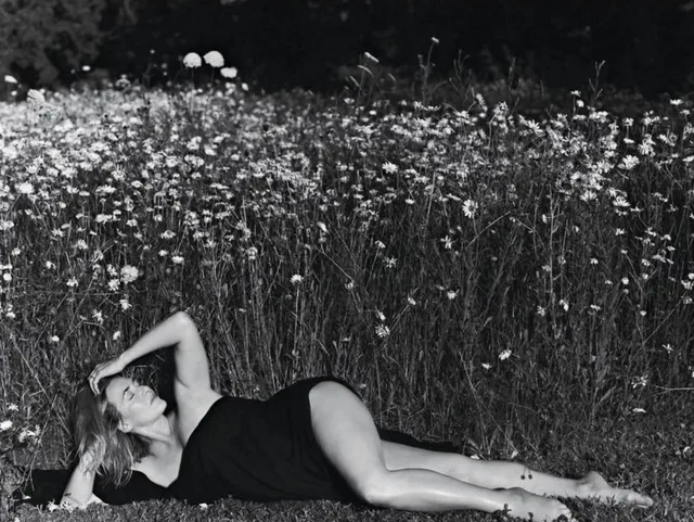 Kate Winslet reclining barefoot in a field in a dark dress.