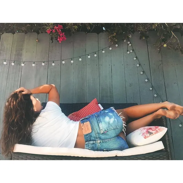 Danielle Campbell reclining in an outdoor bathtub in a white top and shorts.