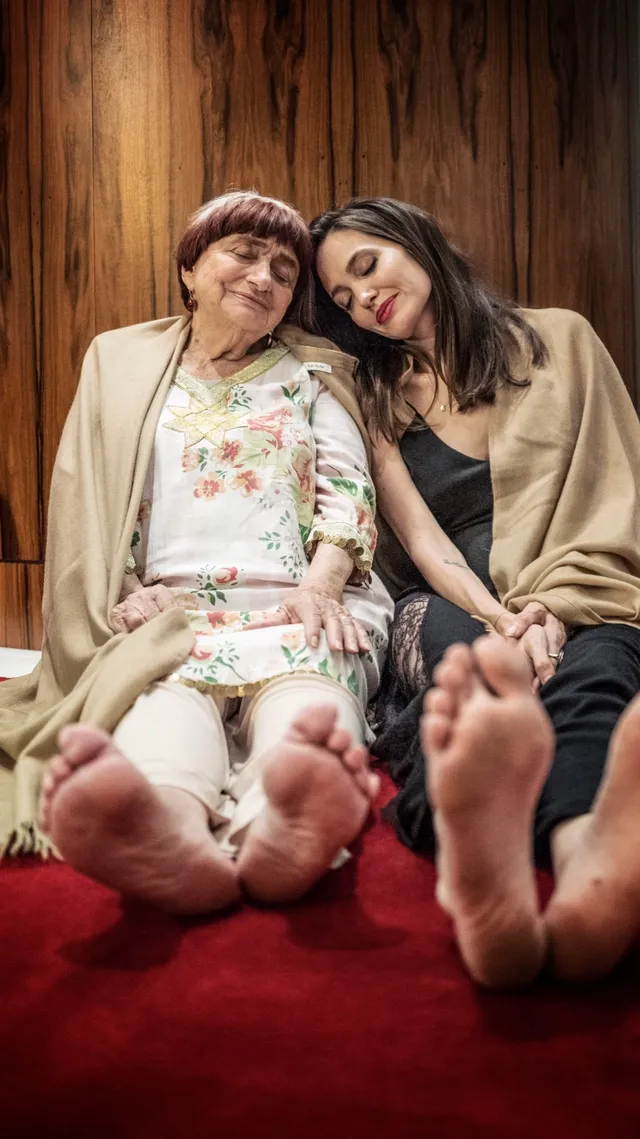 Angelina Jolie seated beside a friend with bare soles raised toward the camera.
