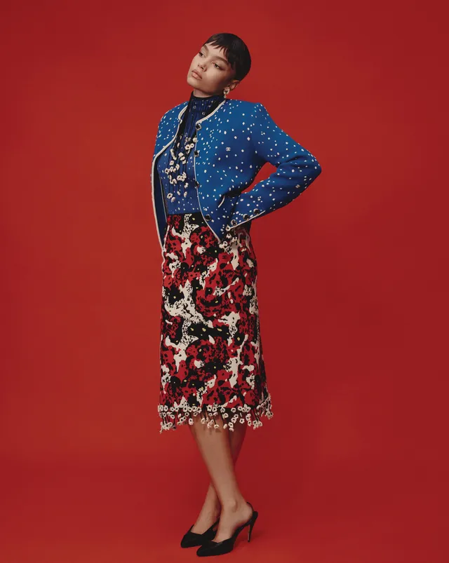 Whitney Peak posing in a blue jacket, patterned skirt and black heels against a red backdrop.