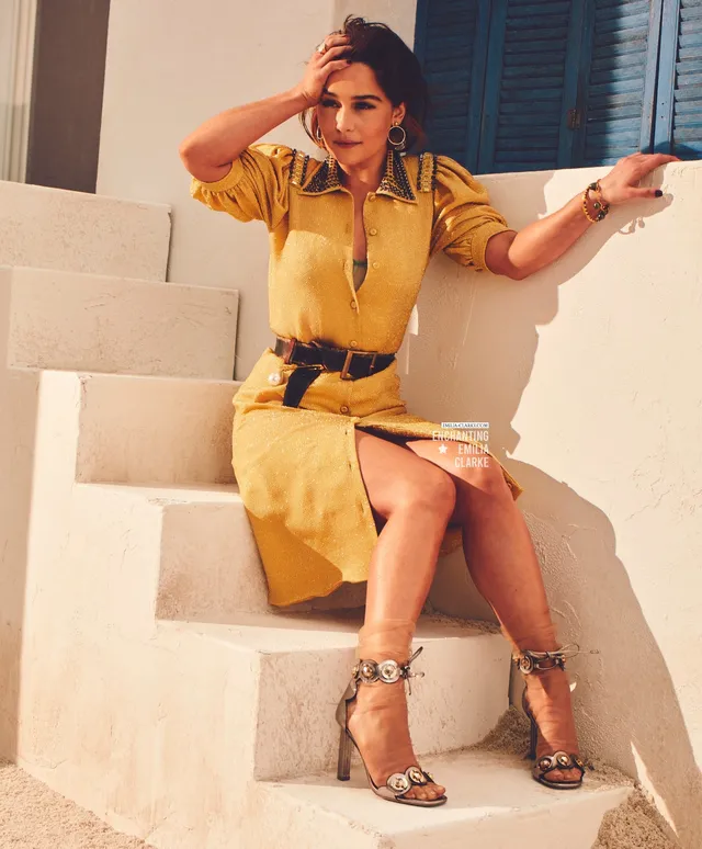Emilia Clarke sitting outdoors in a yellow dress and gold heels.