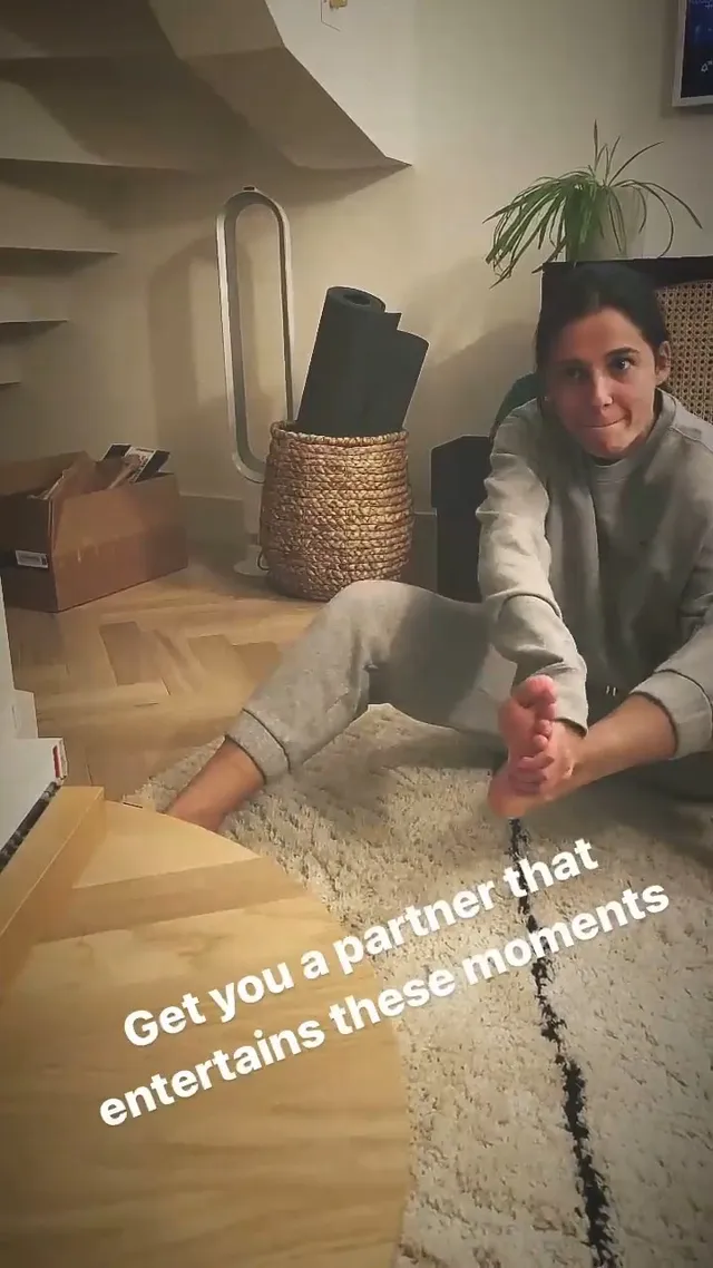 Naomi Scott sitting indoors with a bare foot raised.