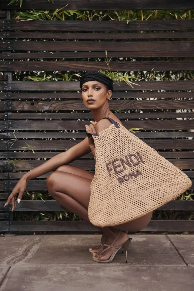 Jasmine Tookes crouching in pale heeled sandals beside a wooden fence.