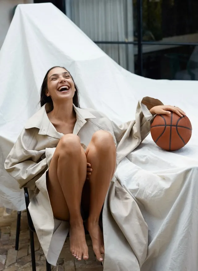 Gal Gadot sitting barefoot in a woven chair with a basketball.