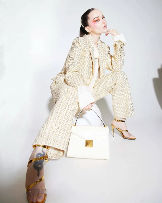 Dove Cameron sitting in a cream suit wearing gold heels.