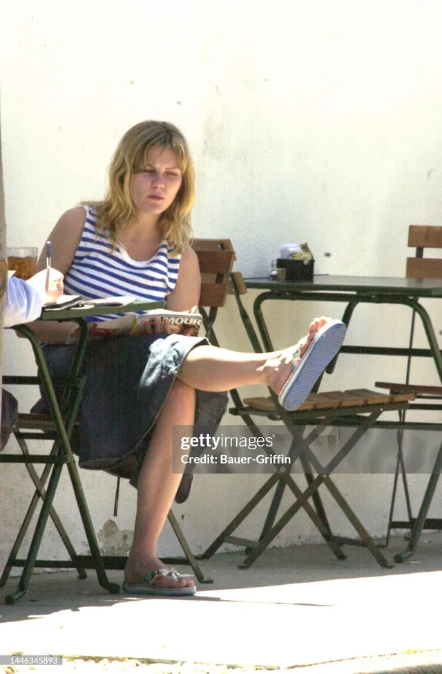 Kirsten Dunst sitting outdoors with one leg extended on a chair.