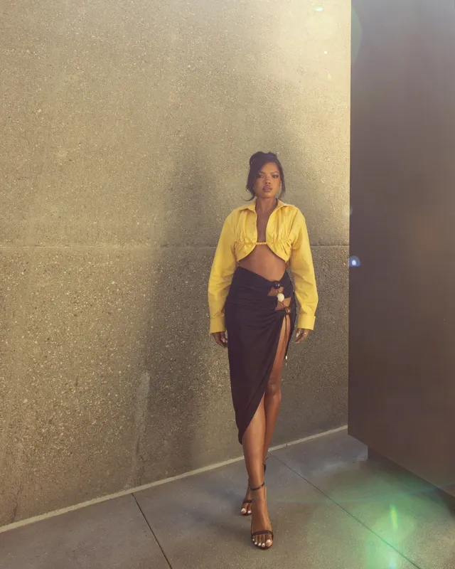 Ryan Destiny posing in a yellow blouse, dark skirt and heels against a wall.