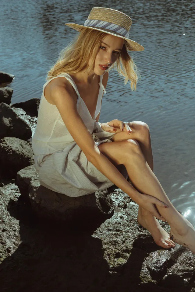 Phoebe Dynevor seated on a rock beside the water in a white dress.