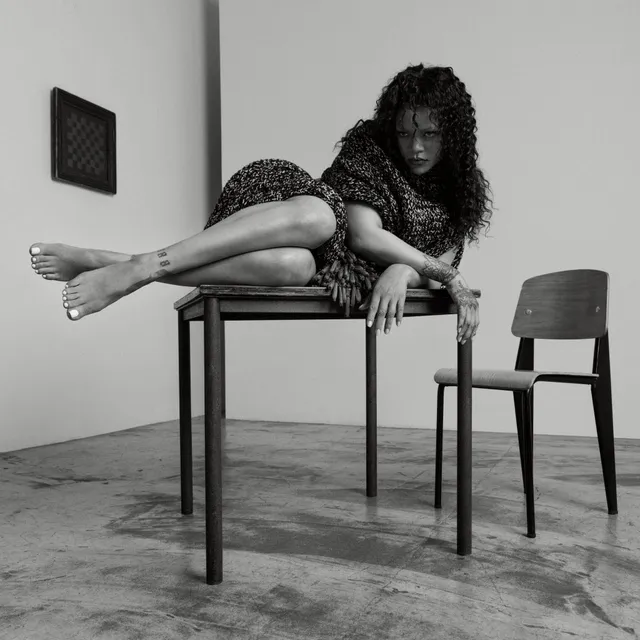 Rihanna sitting barefoot across a wooden table in a monochrome portrait.