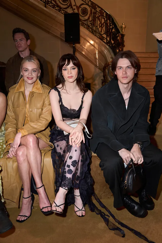 Sophie Thatcher sitting between two people in a patterned dress and heels.