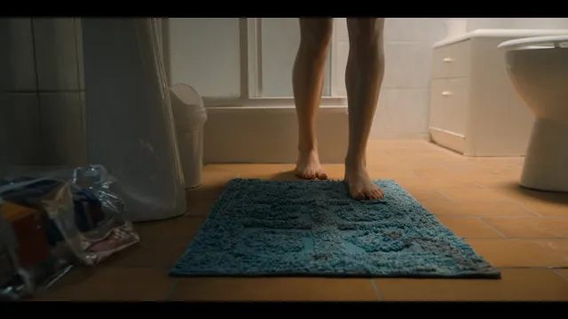 Grace Van Patten standing barefoot on a bathroom mat in a film scene.