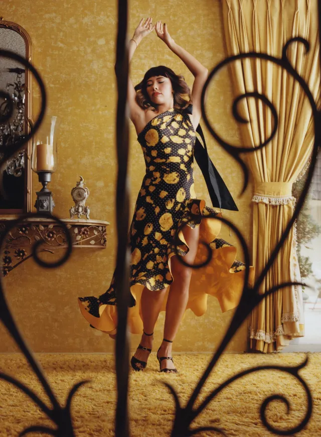 Xochitl Gomez posing in a patterned dress and black heels in a gold room.