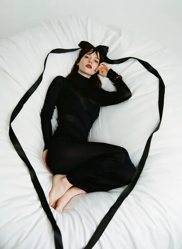 Sophie Thatcher lying barefoot on a white bed in a black outfit.