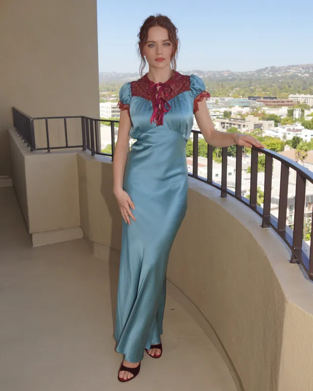 Joey King standing on a balcony in a blue dress and burgundy heels.