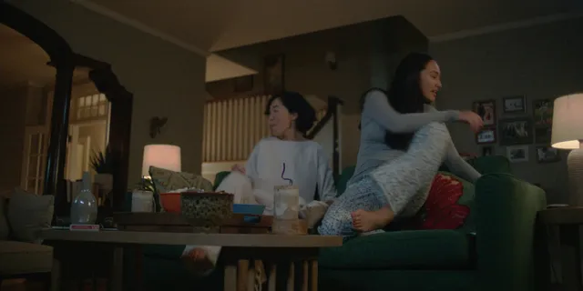 Lola Tung sitting on a sofa with a fellow performer in a scene.