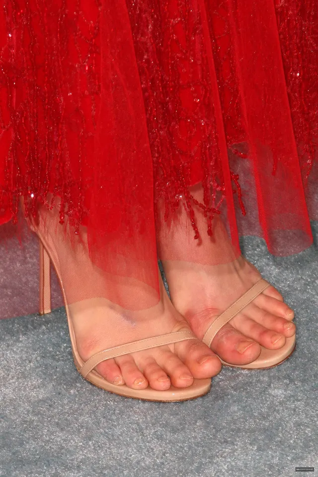 Alexandra Daddario a close view of her feet in pale strappy heels beneath a red dress.