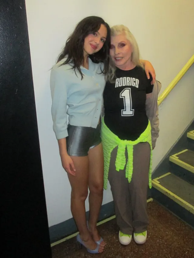 Olivia Rodrigo posing beside another woman wearing a grey top and metallic heels.