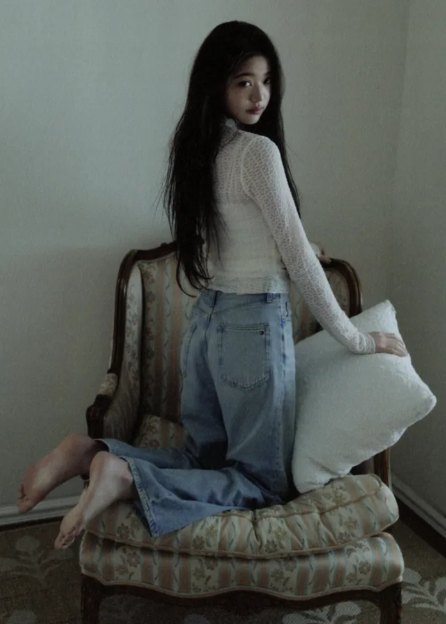 Jang Wonyoung kneeling barefoot on an upholstered chair in jeans.