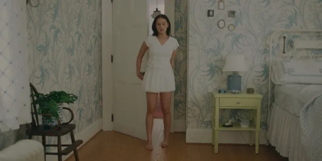 Lola Tung standing barefoot in a white dress in a bedroom scene.