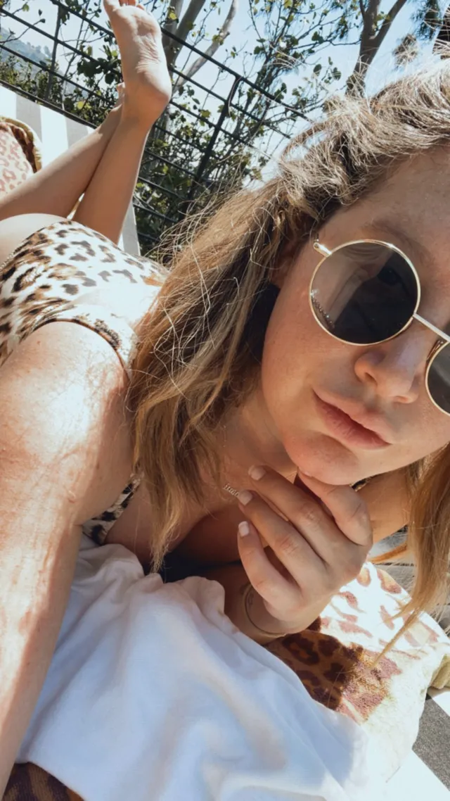 Ashley Tisdale lying barefoot on an outdoor lounger wearing sunglasses.
