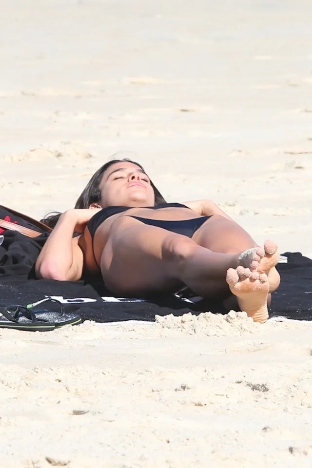 Bruna Marquezine reclining barefoot on a beach towel in a black bikini.