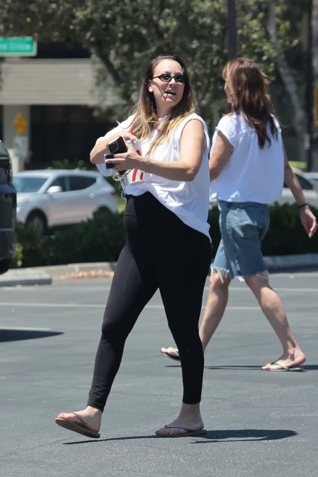 Kaley Cuoco walking outdoors in a white blouse and flip-flops.