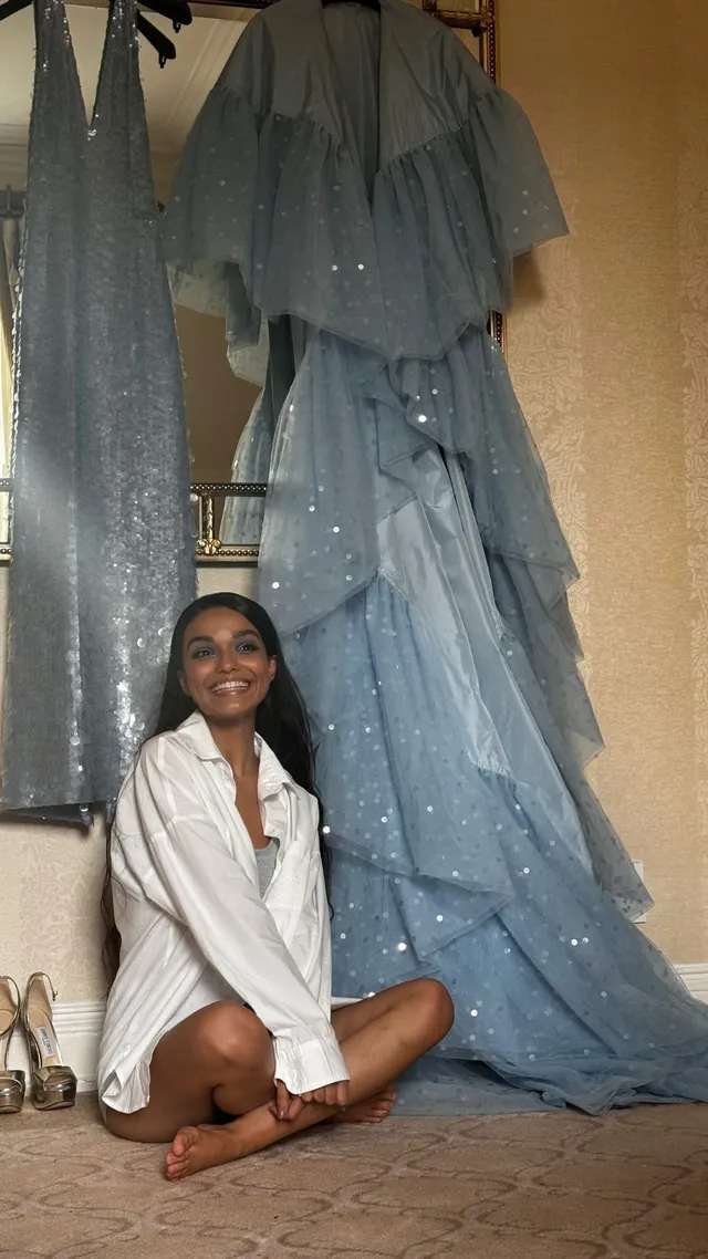 Rachel Zegler sitting barefoot in a white shirt beside hanging gowns.