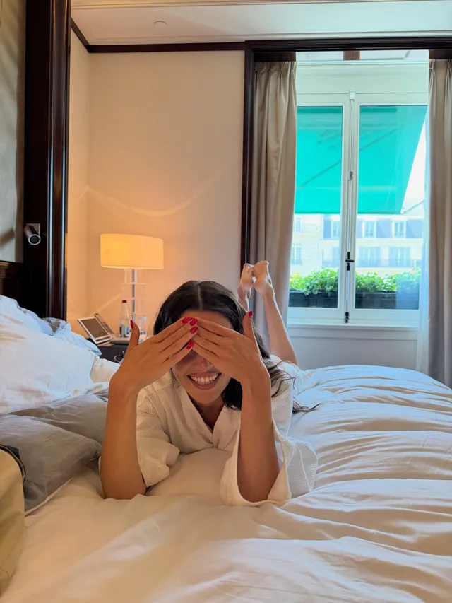 Hande Erçel reclining barefoot on a hotel bed in a white robe.