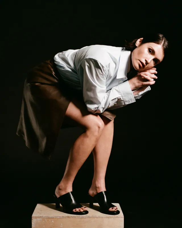 Gemma Arterton sitting on a plinth in a white blouse and black mules.