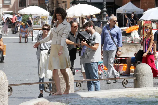 Úrsula Corberó walking barefoot beside a stone fountain in a pale dress.