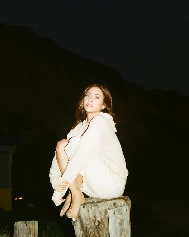 Brittany Snow sitting on a wooden stump in a light outfit.