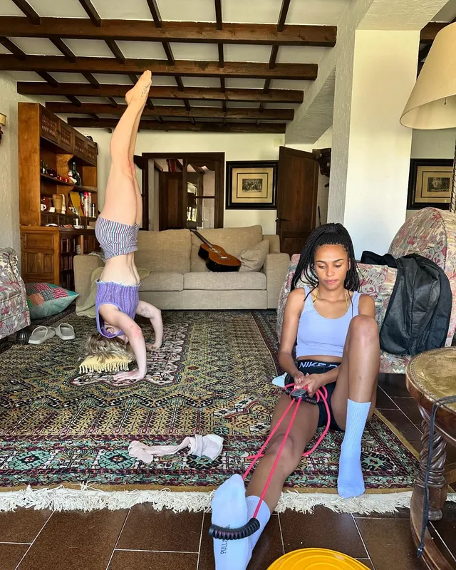 Clara Galle holding a barefoot headstand beside a friend indoors.