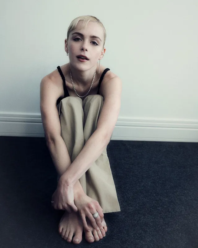 Kiernan Shipka sitting barefoot on a carpet in loose trousers.