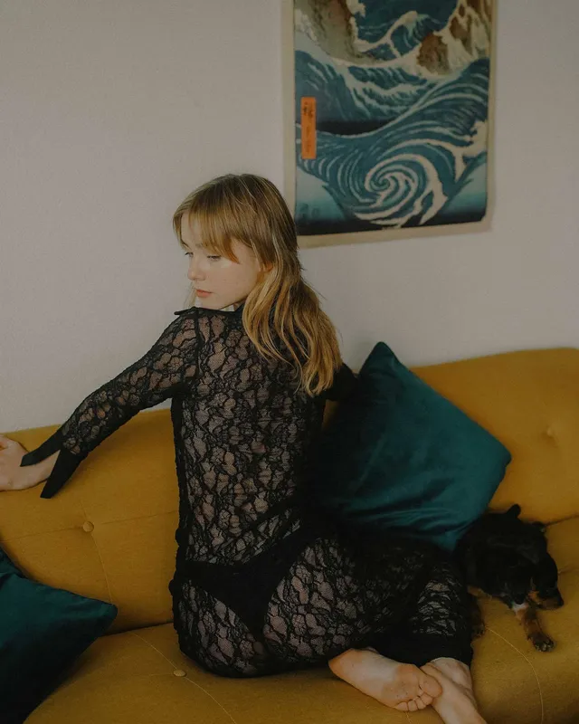 Clara Galle sitting barefoot on a yellow sofa in a black lace dress.