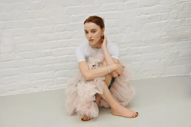 Phoebe Dynevor seated against a white brick wall in a feathered skirt.