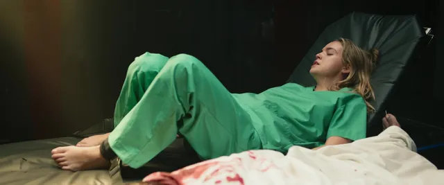 Bailee Madison reclining barefoot in hospital clothing in a scene.