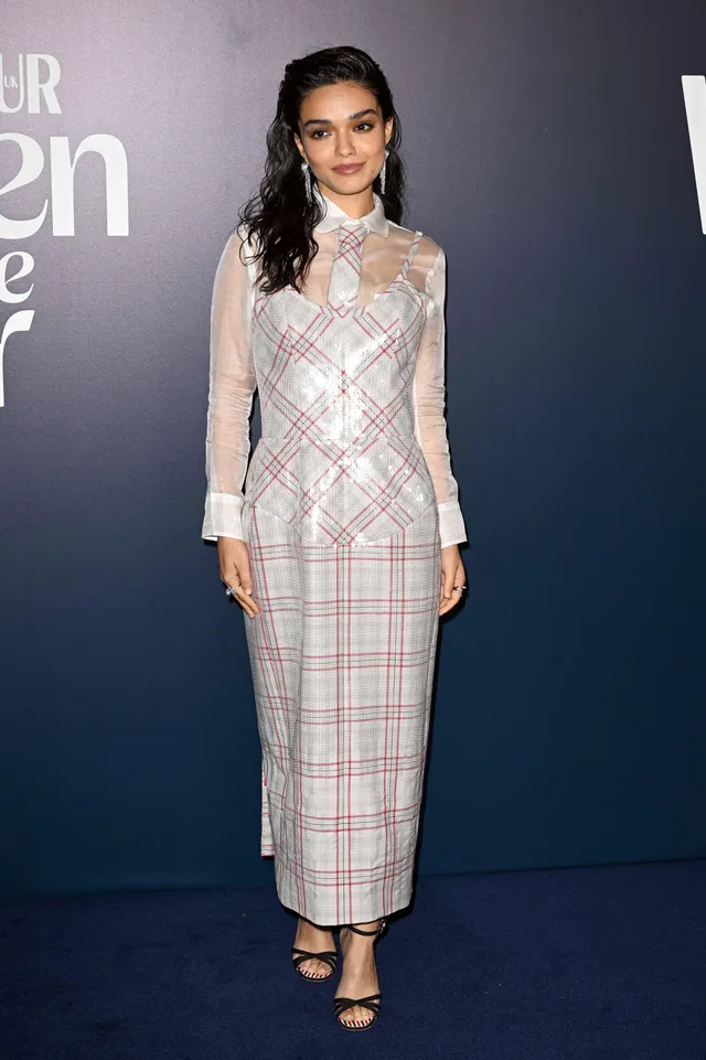 Rachel Zegler standing at an event in a checked dress and black heels.