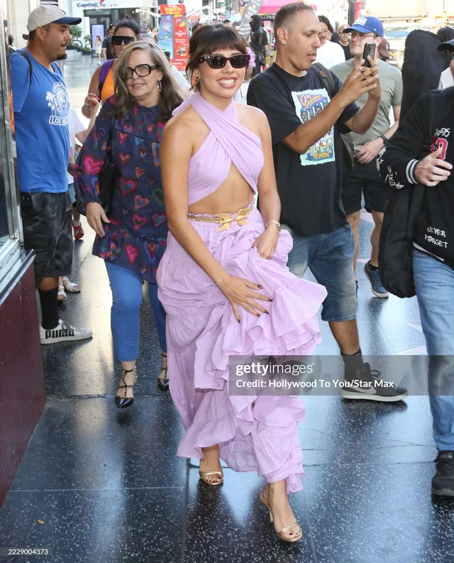 Xochitl Gomez walking outside in a layered purple outfit and pale heels.