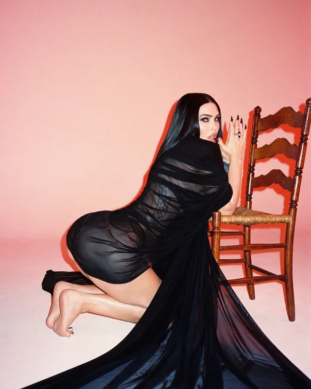 Megan Fox: kneeling barefoot beside a chair against a pink background.