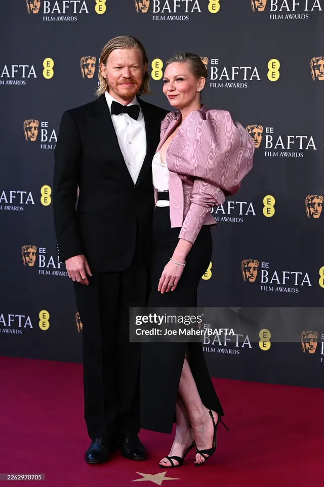 Kirsten Dunst posing with a companion in a pink jacket and black skirt.