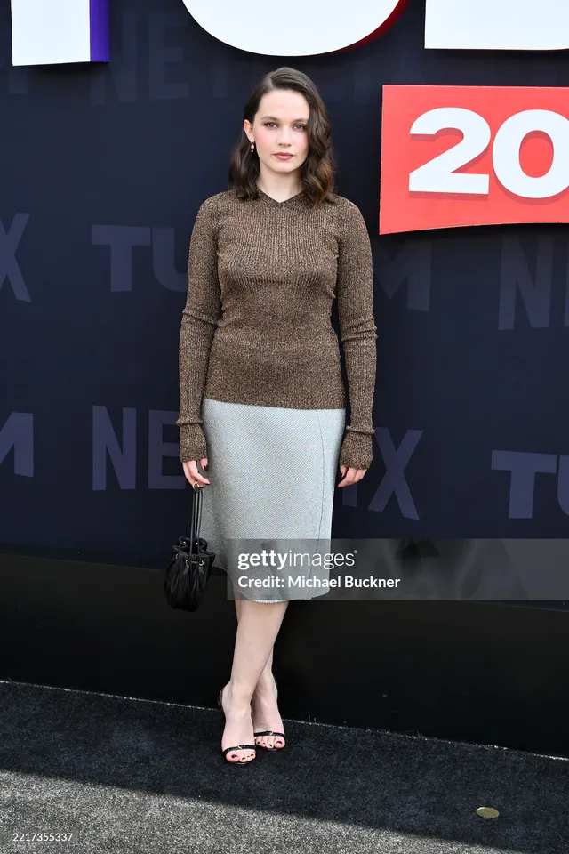Cailee Spaeny posing at a film event in a grey skirt and black heels.