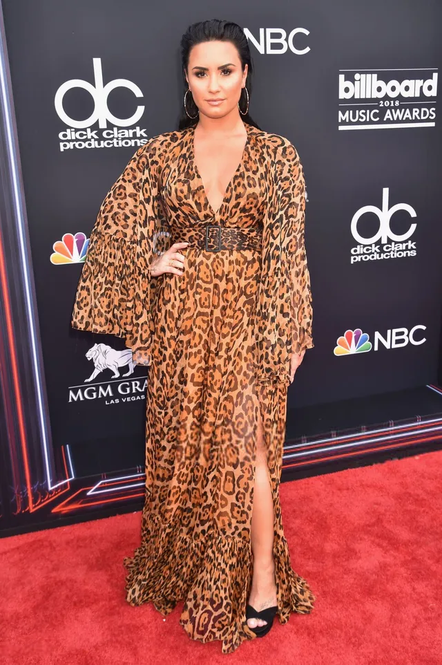 Demi Lovato standing at an event in a patterned gown and black heels.