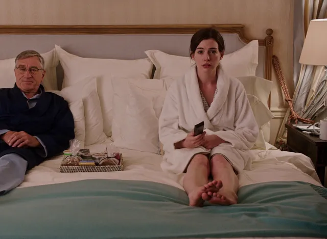 Anne Hathaway sitting on a bed with a companion and bare feet visible.