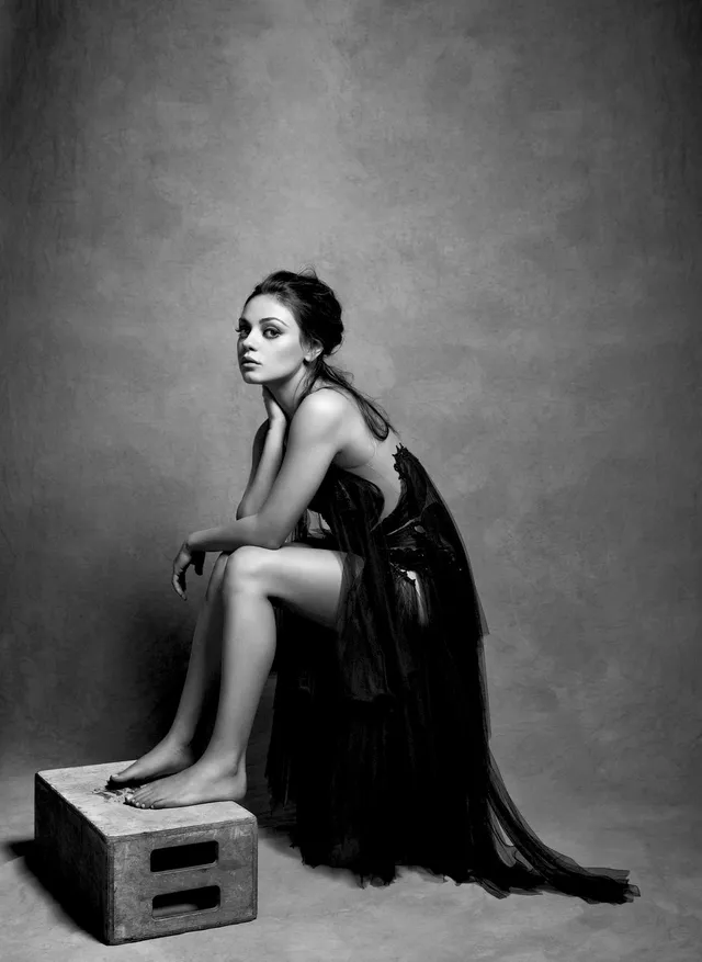 Mila Kunis sitting barefoot on a crate in a monochrome fashion portrait.