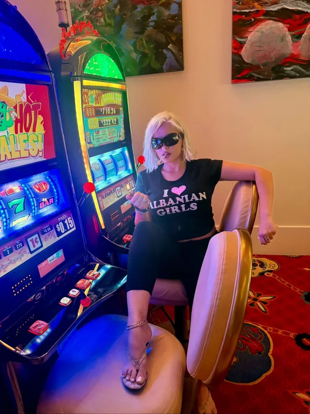 Bebe Rexha sitting beside casino machines wearing sandals.